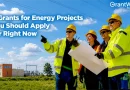 Four utility and energy professionals wearing high-visibility safety gear and hard hats stand at an electrical substation reviewing project plans. Power lines and transmission towers stretch across a bright blue sky in the background. Overlay text reads: "8 Grants for Energy Projects You Should Apply for Right Now," with the GrantWatch logo in the upper-right corner.
