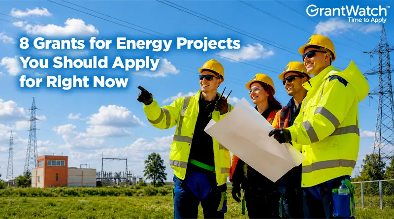 Four utility and energy professionals wearing high-visibility safety gear and hard hats stand at an electrical substation reviewing project plans. Power lines and transmission towers stretch across a bright blue sky in the background. Overlay text reads: "8 Grants for Energy Projects You Should Apply for Right Now," with the GrantWatch logo in the upper-right corner.