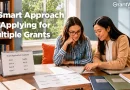 Two women sit at a table reviewing grant application materials and deadlines while planning multiple funding submissions. Documents, folders, and a calendar displaying grant deadlines are spread across the table. A bookshelf and window create a professional office setting. On-screen text reads, “A Smart Approach to Applying for Multiple Grants,” with the GrantWatch logo in the upper-right corner.