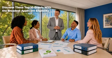 Five professionals sit around a conference table during a collaborative meeting in a bright office with floor-to-ceiling windows overlooking trees and city buildings. A presenter stands at the head of the table speaking to the group. On the table are charts, reports, and binders labeled Education, Nonprofit, Small Business, and Local Government, along with a document titled "Verified Grants." Overlay text reads: "Discover These Top 10 Grants With the Broadest Applicant Eligibility."