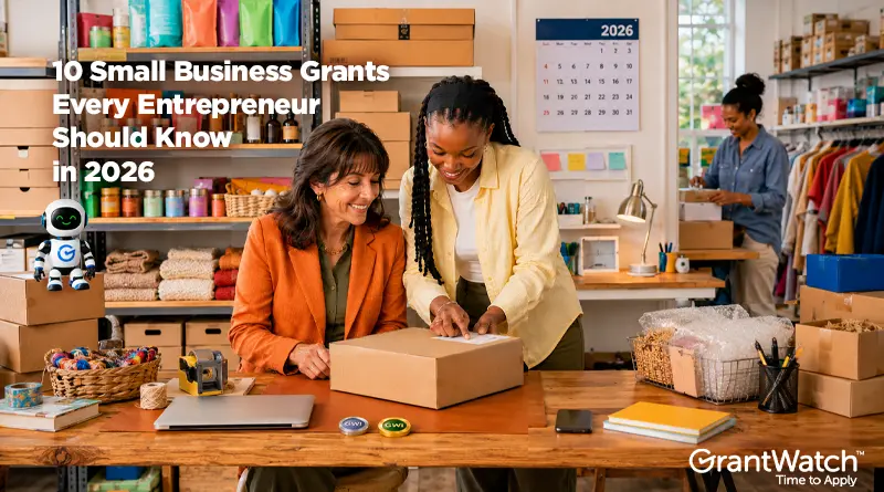 Two women work together in a small business shipping and retail workspace, preparing a package for shipment at a wooden table surrounded by inventory, supplies, and boxes. Shelves stocked with products and a clothing display are visible in the background, while another employee organizes merchandise. A small GrantWatch robot mascot stands on a box to the left. Overlay text reads, "10 Small Business Grants Every Entrepreneur Should Know in 2026." The GrantWatch logo and the tagline "Time to Apply" appear in the lower-right corner.