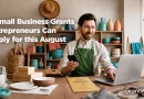 A small business owner sits at a worktable in a pottery studio, holding a smartphone while working on a laptop. Shelves filled with handmade ceramics, tools, and supplies line the background. Text overlay reads: “6 Small Business Grants Entrepreneurs Can Apply for this August.” The GrantWatch logo appears in the bottom right corner.