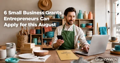 A small business owner sits at a worktable in a pottery studio, holding a smartphone while working on a laptop. Shelves filled with handmade ceramics, tools, and supplies line the background. Text overlay reads: “6 Small Business Grants Entrepreneurs Can Apply for this August.” The GrantWatch logo appears in the bottom right corner.