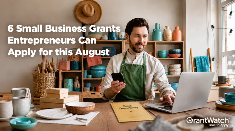 A small business owner sits at a worktable in a pottery studio, holding a smartphone while working on a laptop. Shelves filled with handmade ceramics, tools, and supplies line the background. Text overlay reads: “6 Small Business Grants Entrepreneurs Can Apply for this August.” The GrantWatch logo appears in the bottom right corner.