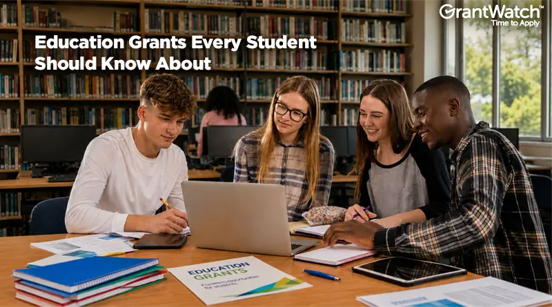 Four students collaborate around a laptop in a library, reviewing notes and educational materials. Bookshelves fill the background, while notebooks, tablets, and a document labeled "Education Grants" sit on the table. The image includes the headline "Education Grants Every Student Should Know About" and the GrantWatch logo in the upper corner.
