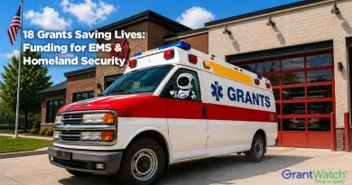 An ambulance labeled "GRANTS" is parked outside a fire station on a sunny day, with an American flag flying nearby. A small robot character is visible inside the ambulance. Text on the image reads: "18 Grants Saving Lives: Funding for EMS & Homeland Security." The GrantWatch logo appears in the lower-right corner.