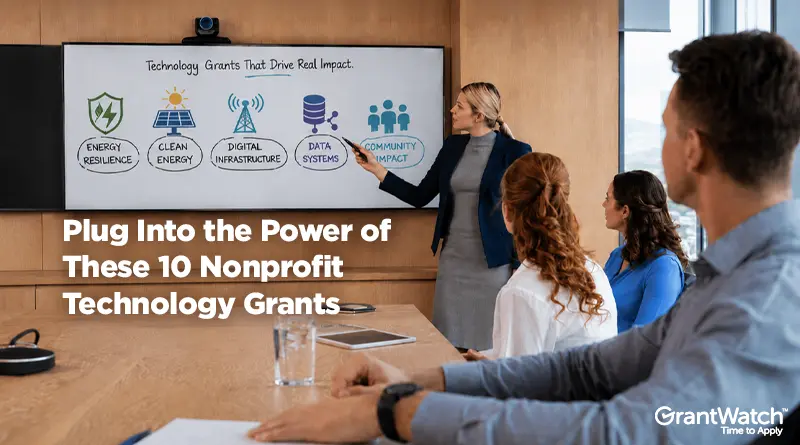 Business meeting in a modern conference room. A presenter stands beside a large display showing a diagram titled “Technology Grants That Drive Real Impact,” with icons representing energy resilience, clean energy, digital infrastructure, data systems, and community impact. Several attendees watch the presentation from around a conference table. Overlaid text reads: “Plug Into the Power of These 10 Nonprofit Technology Grants.” The GrantWatch logo appears in the bottom-right corner.