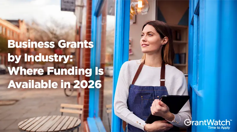 A woman standing in the doorway of a blue storefront, holding a tablet. Large text reads, “Business Grants by Industry: Where Funding Is Available in 2026.” GrantWatch logo appears in the bottom-right corner.